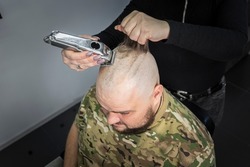 A young man in a military uniform shaves his head bald for military service. A guy with a beard gets a haircut at a barber shop.