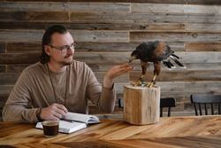 Man is working, writing with wild bird at home by the table. Making noted, memories, diary with eagle as pet. Unusual animals at home. Human friendship. Taking care of buzzard