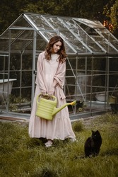 Pretty woman at her 30s in the garden with watering can and cat walking on the grass. Beautiful dreamlike female in the evening walking in long dress over greenhouse. Fashion portrait