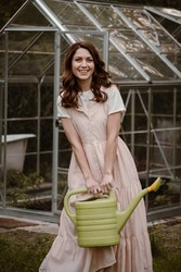 Pretty woman at her 30s in the garden with watering can walking on the grass. Beautiful dreamlike female in the evening walking in long dress over greenhouse. Fashion portrait