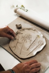 Hands making Clay handmade Ceramic plates, rolled clay on the table with plants and flowers. Top view on handcrafted pottery with floral pattern, close-up