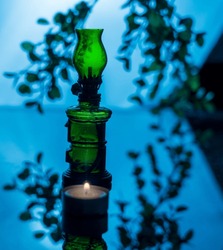 Fairy image of a transparent green glass lamp and a lit candle at its foot, backlit against a background of blurred branches and leaves reflecting on the dark shelf, creating a magical atmosphere