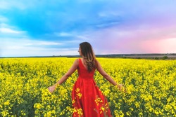 Young beautiful girl in a red dress close up in the middle of yellow field with radish flowers at sunset time