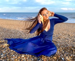 Teenage Girl with a natural long red hair wearing navy blue dress holding seashells on the pebble beach with a sea view