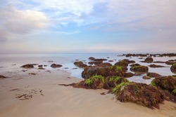 A long exposure shot of rocks on a sandy, pebble beach, shot at dusk with sunset colours, rich colours, movement in clouds, seaweed covered rocks and space for text
