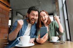 Overjoyed couple of friends holding mobile phone shopping online with sales, win money, celebration success sitting in modern cafe. Happy successful freelancers receive payment, reading good news 