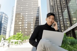 Portrait of handsome asian man using laptop, talking on mobile phone, outdoors. Young chinese student preparing for exam, sitting on city street