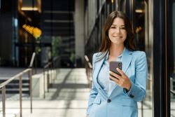 Portrait of beautiful smiling businesswoman wearing formal wear, business suit, holding phone, texting message, looking at camera, copy space. Attractive female online shopping, mobile banking