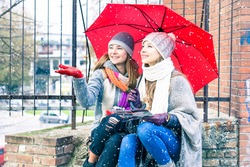 Young women reaching snowflakes under red umbrella at winter holiday - Happy female friends sitting at street corner holding city tour map looking up at snowy weather - Forecast and vacation concept