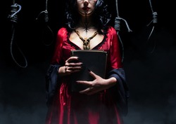 Witch or sorceress with runic makeup and wooden animal skull amulet holding a magic book on dark room background with nooses. Halloween concept.