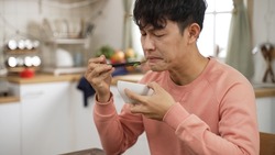 asian Japanese man grimacing face while trying a piece of vegetable in dining room at home. he finds the food too salty and puts down chopsticks