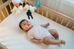 adorable asian newborn child is looking upward at the colorful hanging toys with curiosity while playing alone in the baby bed with white bedding at home.