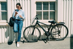 full length asian female is using mobile phone by her bike at leisure while standing against the wall of a white tin house with coffee in the sun at summer time