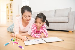 Mother and daughter are enjoy drawing and girl learning how to pain on notebook lying down on wooden floor having fun while drawing in the living room at home. family activity concept.