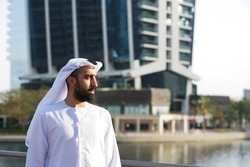 Handsome Arab Emirati businessman in UAE looking to side in summer sunlight wearing traditional dress outdoors looking ambitious and confident
