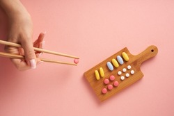 Women's hands hold pill with chopsticks on minimal pink background. Creative concept photo with variety of nutritional supplement vitamins or pills arranged as sushi plate. 