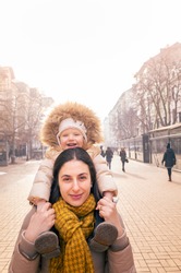 Cute young happy two year old girl on the shoulders of her mother outdoors on a city street on a cold winter day	