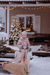 Happy cute girl in winter clothes tying a bow on a gift sitting on a snow-covered porch of a house decorated with holiday decorations, a Christmas tree and a garland. Waiting for Christmas.