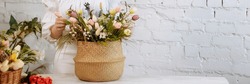A young woman florist in white clothes and a straw hat stands with a basket of flowers against a white brick wall, in a flower shop. Banner for website header design with copy space.