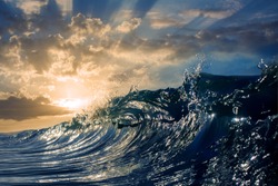 An ocean sunset shorebreak in side view with bokeh flares. Big beautiful high contrast sunrise wave swirling. Bright sun shining on blue sky with colorful clouds.