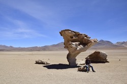 A man is sitting on a rock next to the famous stone tree rock formation (Arbol de Piedra) in the Siloli desert in the region of the Uyuni Salt Flat, Bolivia, South America.