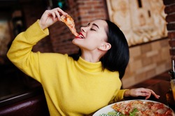 Funny brunette girl in yellow sweater eating pizza at restaurant. 