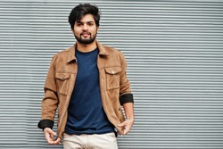Stylish casual indian man wear blue t-shirt and brown jacket posing against grey wall.