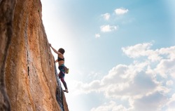A woman climbs a rock against a blue sky, a strong girl trains strength and endurance, an extreme sport, rock climbing on natural terrain, a rock climber climbs with a rope