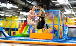 Pretty girls jumping together on colorful trampoline at playground park. Two sisters having fun during active entertaiments indoor