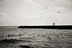 Art photo. A couple in love stands on a pier by the sea with waves, seagulls fly around in the sky with clouds. Black and white silhouette photography.