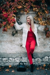 Portrait of a positive smiling woman with blond long hair and stylish make-up with red lips, in a light winter coat, against the background of vine branches, near a concrete wall, on a city street.