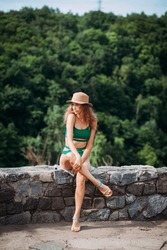 A slender positive girl with blond hair, tanned skin, dressed in a green stylish swimsuit and a hat, sits against the backdrop of a picturesque landscape with forests, a river and a waterfall.