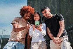 Young multicultural teenagers watching funny images on a smart phone. Three smiling young adult people browsing on internet with a celphone app. Hppy students playing video games using a mobilephone