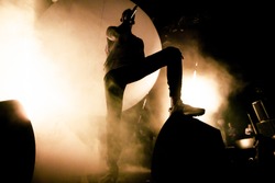 A Singer is on the stage. A silhouette of the singer is putting his foot on a speaker. A brutal shadow of a rapper on the stage. Smoke and bright soft yellow stage lights in the background.