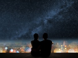 Silhouette of young Asian couple sit on wooden ground above the city under stars.