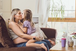 pregnant mom and little toddler daughter baby girl kissing hugging on sofa living room home