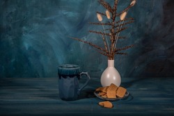 Gingerbread cookies on a saucer, a cup of tea and a bouquet of dry branches in a vase on a dark blue background.
