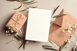 Greeting card mockup with gift box, envelope and eucalyptus and gypsophila twigs on beige background. Card mockup with copy space.