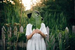 Stylish girl in linen dress holding big green leaf at face at wooden fence and grass. Portrait of boho woman in hat posing with leaf in summer countryside in evening. Atmospheric moment
