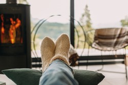 Feet in warm knitted woolen socks on background of modern black fireplace and big windows with view on mountains. Woman relaxing in comfortable home, cozy warm moments
