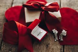 Happy Valentines day. Stylish gift box with red ribbon, luxury pearl ring and earrings with stones on rustic wooden background with red velvet hearts. Love and marriage concept