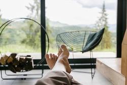Beautiful female feet on background of firewood and big window in modern chalet. Young woman barefoot relaxing in comfortable home with amazing view on mountain hills, cozy leisure time