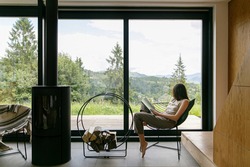 Beautiful stylish woman reading book on chair at fireplace with firewood on background of mountain hills. Young female in casual clothes relaxing in modern chalet with amazing view from window