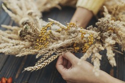 Hands making stylish autumn rustic wreath with dry grass, wildflowers and wheat on rustic wooden table in room. Fall holiday workshop. Florist making boho wreath on dark wood