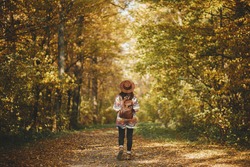Stylish woman hipster with backpack in hat walking in sunny autumn woods. Young female traveler hiking in fall forest, beautiful moment. Travel and wanderlust concept, space for text. Back view