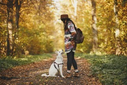 Stylish woman traveler in hat hiking with cute dog in sunny autumn woods. Young female hipster traveling with swiss shepherd white dog. Travel with pet, loyal companion. Wanderlust
