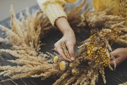 Making stylish autumn wreath. Hands holding dry grass, wildflowers and wheat and making rustic wreath on wooden table. Fall holiday workshop. Florist making boho wreath on dark wood