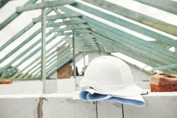 Hard hat with house blueprints on aerated concrete blocks on background of unfinished wooden roof framing. Architecture and House building concept. Safety helmet on construction site