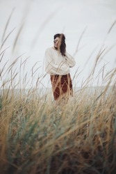 Beautiful stylish woman with windy hair in knitted sweater posing among wild grass. Carefree moment, stylish image. Fashionable young female standing on windy coast. Authentic and Tranquility