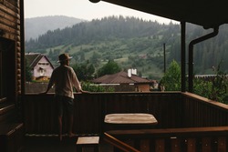 hipster man standing on porch of wooden house and relaxing looking at mountains in evening, calm moment, summer vacation concept, space for text